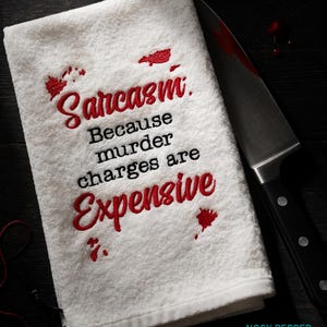 May include: White towel with the embroidered text "Sarcasm. Because murder charges are Expensive" in red and black. A large knife with a black handle and a blood-like stain is next to the towel.