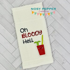 Peut inclure: Serviette de toilette blanc cassé avec la phrase brodée "Oh Bloody Hell" en noir et rouge. Un cocktail Bloody Mary de dessin animé est brodé en dessous du texte, avec une tige de céleri verte et un visage souriant.