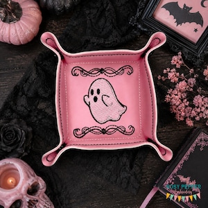 May include: A square pink valet tray featuring a black embroidered ghost design in the center, flanked by decorative black scrollwork borders. The tray has raised, snap-button corners and is constructed with visible black stitching along the edges.