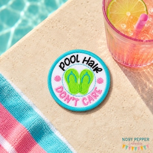 Könnte beinhalten: Gestickter Aufnäher mit dem Text "Pool Hair Don't Care" in Schwarz und Rosa. Der Aufnäher zeigt ein Paar grüne Flip-Flops in der Mitte, umgeben von einem türkisfarbenen Rand und kleinen Muschelmotiven. Ein erfrischendes Getränk mit einer Limettenscheibe ist im Hintergrund.
