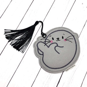 May include: Grey felt bookmark shaped like a seal with pink cheeks and a black tassel. The bookmark has the text "Polka Dot Creations" on the bottom.