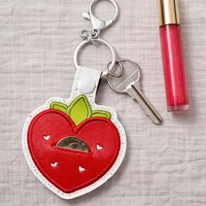 May include: A red strawberry-shaped keychain with a white border and green leaves. The keychain has a small pocket with a coin inside. A silver key and a tube of red lip gloss are next to the keychain.