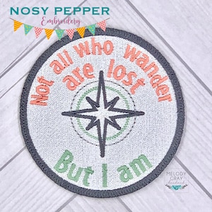 May include: A round, white embroidered patch with a compass design and the text "Not all who wander are lost, but I am". The patch has a gray border and is on a white wooden background.