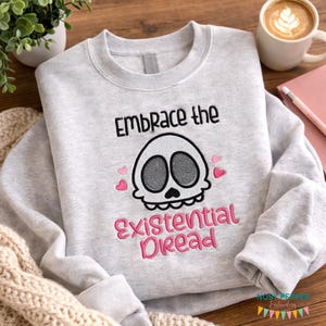 May include: A light gray sweatshirt with the embroidered text "Embrace the Existential Dread" above a skull design with pink hearts. The skull has gray eyes and a black mouth. The sweatshirt is on a wooden surface.