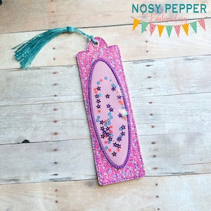 May include: A pink glitter bookmark with a turquoise tassel. The bookmark features a central oval filled with colorful star-shaped confetti. The text "NOSY PEPPER Embroidery" is at the top.