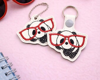 May include: Two white panda keychains with red glasses, black eyes, and black accents. The keychains are on a pink surface with red sunglasses, a pink lip gloss, and a blue spiral notebook. The keychains are embroidered.