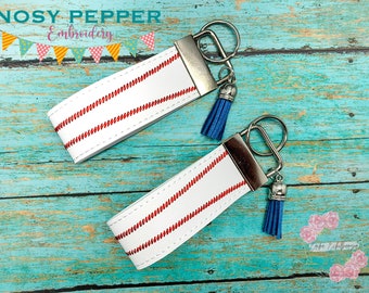 May include: Two white leather keychains with red stitching, resembling a baseball, attached to silver key rings with blue tassels. The keychains are on a turquoise wooden background.