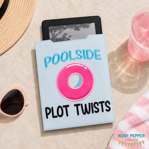 May include: A light blue e-reader case with the words "Poolside Plot Twists" embroidered in turquoise and black. A pink inflatable ring is embroidered in the center. A black e-reader is inside the case. Sunglasses, a hat, and a drink are in the background.