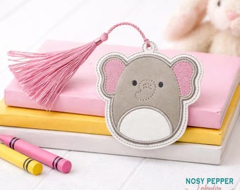 May include: A gray elephant-shaped bookmark with pink ears and a white belly, attached to a pink tassel. The bookmark is on top of a stack of books with a pink and yellow cover. The image also includes two crayons.