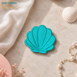 Op de afbeelding: Een turquoise schelpvormige geborduurde patch. De patch ligt op een neutrale ondergrond met een witte zeeschelp en een parelketting. De merknaam "NOSY PEPPER" staat linksboven.