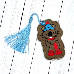 Peut inclure: Un marque-page en feutre marron avec une image brodée d'une marmotte portant un chapeau bleu, une écharpe rouge et des bottes rouges. Le marque-page a un pompon bleu.