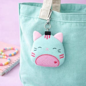 Puede incluir: Un llavero con forma de gato azul claro con detalles en rosa, sujeto a un bolso tote azul claro. El llavero presenta detalles bordados, incluyendo una barriga rosa, orejas rosas y un pequeño corazón rojo. El llavero está sujeto con un cierre plateado.