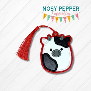May include: A red and white cow bookmark with a black and white face and a red tassel. The bookmark is embroidered with a black outline.