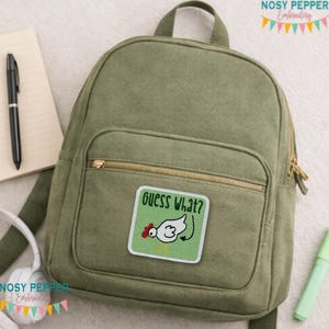 May include: Olive green backpack with a gold zipper and a patch that says "Guess What?" with a cartoon chicken design. The backpack has a top handle and adjustable straps. A notebook, pen, and headphones are visible in the background.
