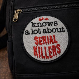 Puede incluir: Mochila negra con un parche redondo que dice "knows a lot about SERIAL KILLERS" en bordado negro y rojo. La bolsa tiene una cremallera dorada y un bolsillo pequeño.