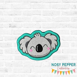 May include: An embroidered patch featuring a smiling koala bear with gray fur, black eyes, and pink cheeks, outlined in teal. The patch has the text "NOSY PEPPER Embroidery" below it, with a banner of colorful flags.