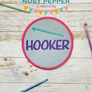 May include: A round, gray embroidered patch with a pink border. The patch features a turquoise crochet hook and the word "HOOKER" in purple lettering. The patch is on a white wooden background.