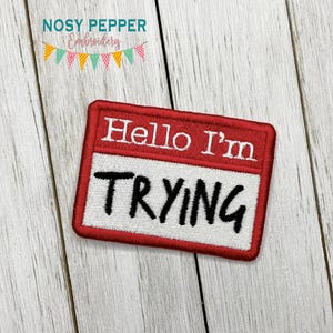 以下が含まれることがあります： 「Hello I'm TRYING」という黒い文字が書かれた、赤と白の刺繍入りアイロン接着パッチです。