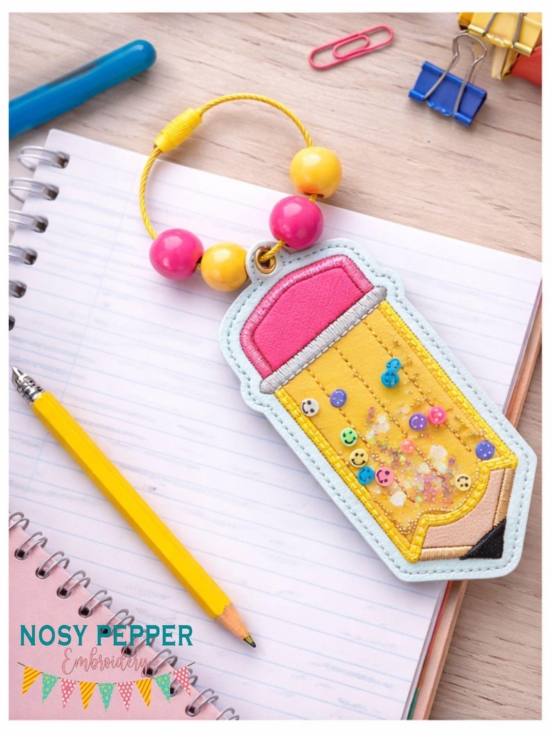 May include: A yellow pencil-shaped keyring with a pink eraser and a light blue border. The keyring has a yellow beaded loop with pink and yellow beads. The pencil design includes small smiley face embellishments and the text "Nosy Pepper Embroidery".