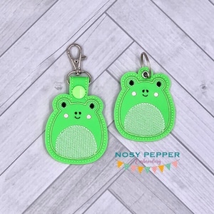 May include: Two green frog-shaped keychains with embroidered details. One keychain has a metal ring and a clip, while the other has a metal ring and a loop. Both keychains feature a smiling frog face with white eyes and a white belly.