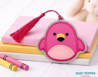 May include: A pink penguin bookmark with a red tassel, resting on a stack of books. The penguin has a light pink belly, black eyes, and a yellow beak. The bookmark is next to yellow and pink crayons.