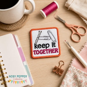 Puede incluir: Un parche bordado cuadrado con borde naranja que muestra una horquilla, un clip y un imperdible sobre el texto negro y rosa "keep it TOGETHER". El parche está rodeado de artículos de costura, incluyendo hilo, tijeras y un descosedor.