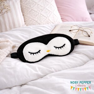 Puede incluir: Una máscara para dormir con forma de pingüino en blanco y negro con los ojos cerrados y un pico amarillo descansa sobre una cama blanca. La máscara tiene una textura suave y afelpada y una banda elástica negra. El fondo incluye almohadas, un libro y gafas.
