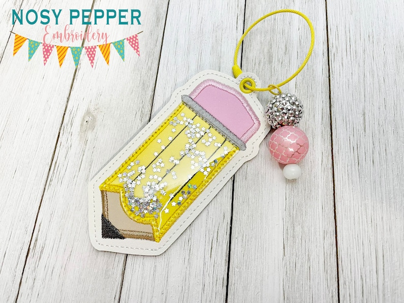 May include: A yellow pencil-shaped bookmark with a pink eraser and silver glitter inside. The bookmark has a yellow loop and is adorned with decorative beads. The top left corner of the image has the text "NOSY PEPPER Embroidery".