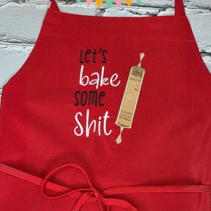 May include: A red apron featuring the embroidered text "Let's bake some shit" and a rolling pin design. The apron has a neck strap and waist ties. The apron is made of a solid red fabric.