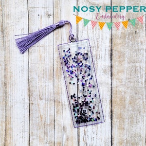 May include: A clear bookmark with a purple tassel. The bookmark is filled with iridescent glitter and stars. The bookmark is on a light wood background.