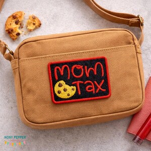 Peut inclure: Sac bandoulière en toile marron avec un écusson rectangulaire « MOM TAX » en rouge et jaune. L'écusson présente un motif de biscuit. Un cookie aux pépites de chocolat cassé est visible en haut à gauche.