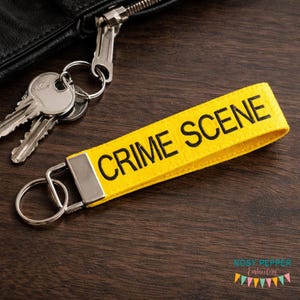 May include: A yellow fabric keychain with the words "CRIME SCENE" in black lettering. The keychain is attached to a silver metal ring and is next to a set of keys and a black leather wallet.