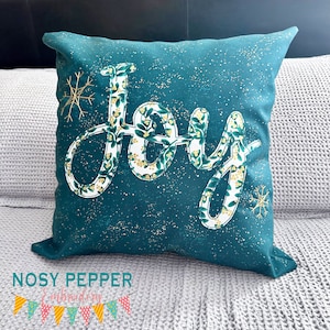 May include: A teal blue throw pillow with a white "Joy" design featuring a green floral pattern. The pillow has a subtle gold glitter effect.