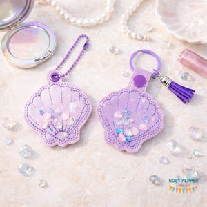 Puede incluir: Dos llaveros en forma de concha en tonos lavanda. Cada llavero presenta un contorno cosido, relleno de confeti iridiscente y una borla a juego. Uno tiene una cadena, el otro un anillo.