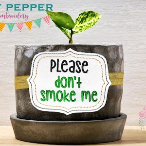 May include: A small potted plant with a white sign that says "Please don't smoke me". The sign is decorated with gold stitching. The plant is in a grey pot with a gold ribbon around it.