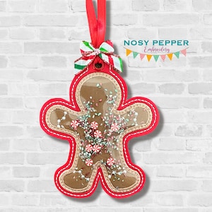 May include: A red felt gingerbread man ornament with a clear plastic window showing sprinkles and candy canes inside. The ornament has a red ribbon and a white and green bow.