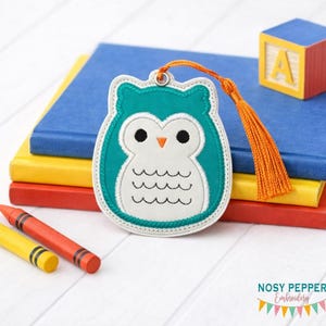 May include: An owl-shaped bookmark with a teal body, white face, and orange tassel. The bookmark is embroidered with black details and a stitched border. It rests on a stack of colorful books and is next to a wooden block.