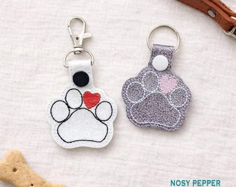 May include: Two paw-shaped keychains. One is white with a red heart and black outline, the other is gray with a pink heart. Both have a metal clasp and ring. A dog bone biscuit is in the lower left corner.