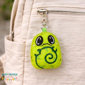 May include: A lime green plush keychain with a cartoon-like face and a green embroidered design. The keychain is attached to a silver zipper pull on a beige backpack. The keychain has black eyes and a smiling mouth.
