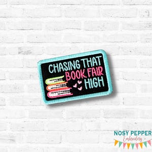 May include: Embroidered patch with the text "CHASING THAT BOOK FAIR HIGH" in white and pink on a black background. The patch features three stacked books and pink hearts, all outlined in a light blue border. The patch is rectangular.