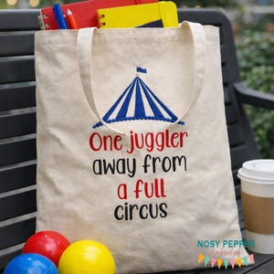 May include: A beige canvas tote bag with a blue and white circus tent design and the text "One juggler away from a full circus" in red and black. The bag is filled with notebooks and pens. Colorful juggling balls are in the foreground.