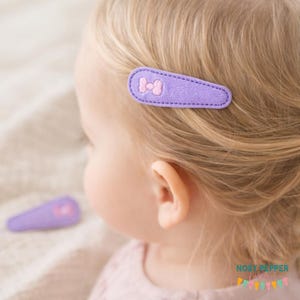 May include: A close-up of a child's head with a purple hair clip. The clip is rectangular with a stitched border and a pink bow. A second clip is visible in the background. The child has blonde hair.
