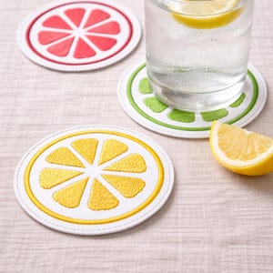 May include: Embroidered fruit slice coasters in red, green, and yellow. A glass of water with a lemon slice sits on a green coaster. A lemon wedge rests nearby. The coasters have a white border.