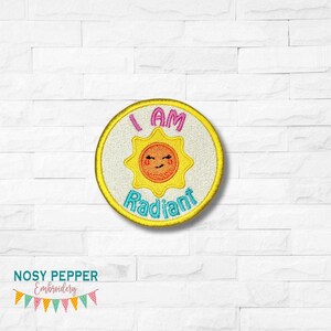 Può includere: Un cerotto rotondo bianco ricamato con un sole giallo con un viso sorridente e il testo "I AM RADIANT" in rosa, blu e giallo.