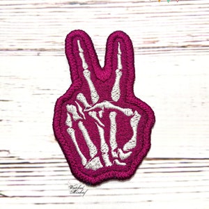 May include: A pink embroidered patch of a skeleton hand making the peace sign.