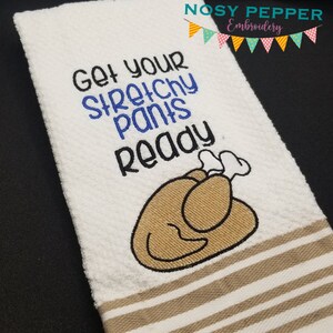 May include: White kitchen towel with the embroidered text "Get your STRETCHY PANTS Ready" in black and blue. A cartoon turkey is embroidered below the text. The towel has tan stripes at the bottom.