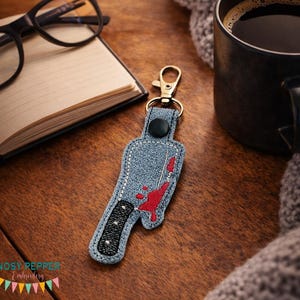 May include: A denim-textured keychain featuring an embroidered knife with red blood splatter. The keychain has a black handle and a gold-colored clasp. The text "Nosy Pepper Embroidery" is visible at the bottom.