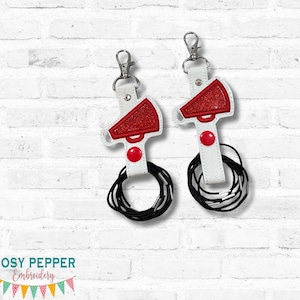 May include: Two white and red megaphone keychains with black hair ties. The megaphone tops are red with glitter, and the keychains have silver clasps. The keychains are on a white brick background.