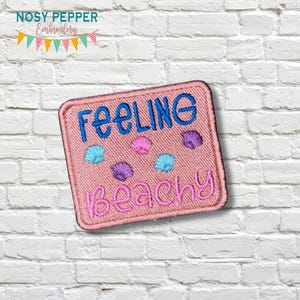 May include: A pink embroidered patch with the words "Feeling Beachy" in blue and pink. The patch features seashell designs in blue, pink, and purple. The patch is square-shaped with a pink border. The brand name "Nosy Pepper Embroidery" is at the top.