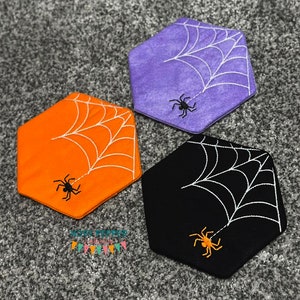 May include: Three hexagon-shaped coasters with a spider web and spider design. The coasters are orange, purple, and black.